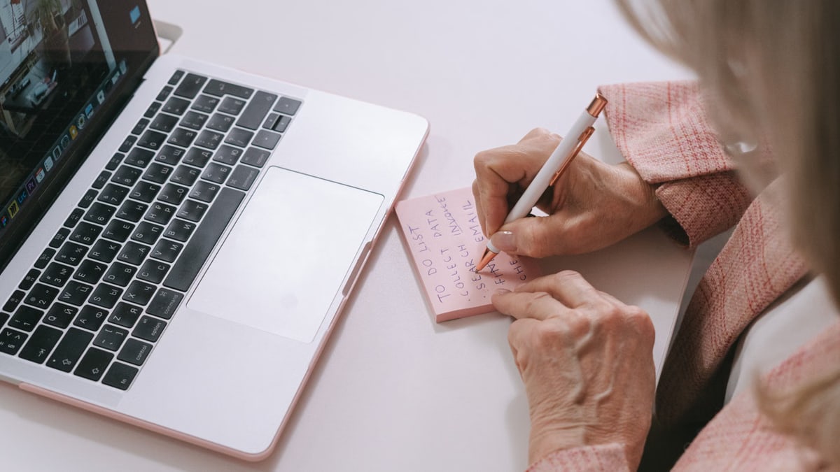 Close-up of a woman writing on a pink sticky note beside a laptop. Productive work setting.